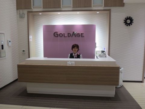 受付スタッフである。女性が笑顔で座っており、首元にはスカーフを巻いている。背面には紫が使われており、落ち着いた雰囲気がある。