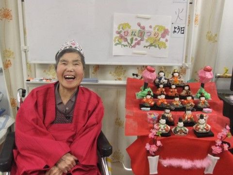 ひな祭りの日に、ひな壇の横で口を開けて笑顔を見せる女性入居者様