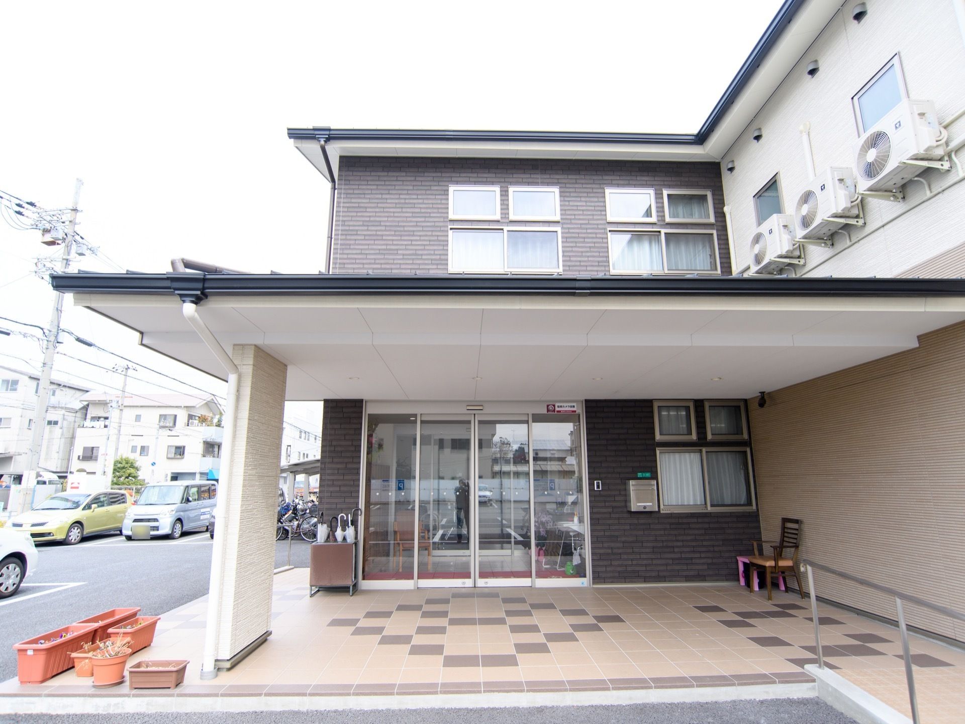 広々とした玄関前スペースである。大きな屋根があり、ガラス張りの玄関ドアの隣に、傘立てを置いている。横側の壁際に椅子がある。