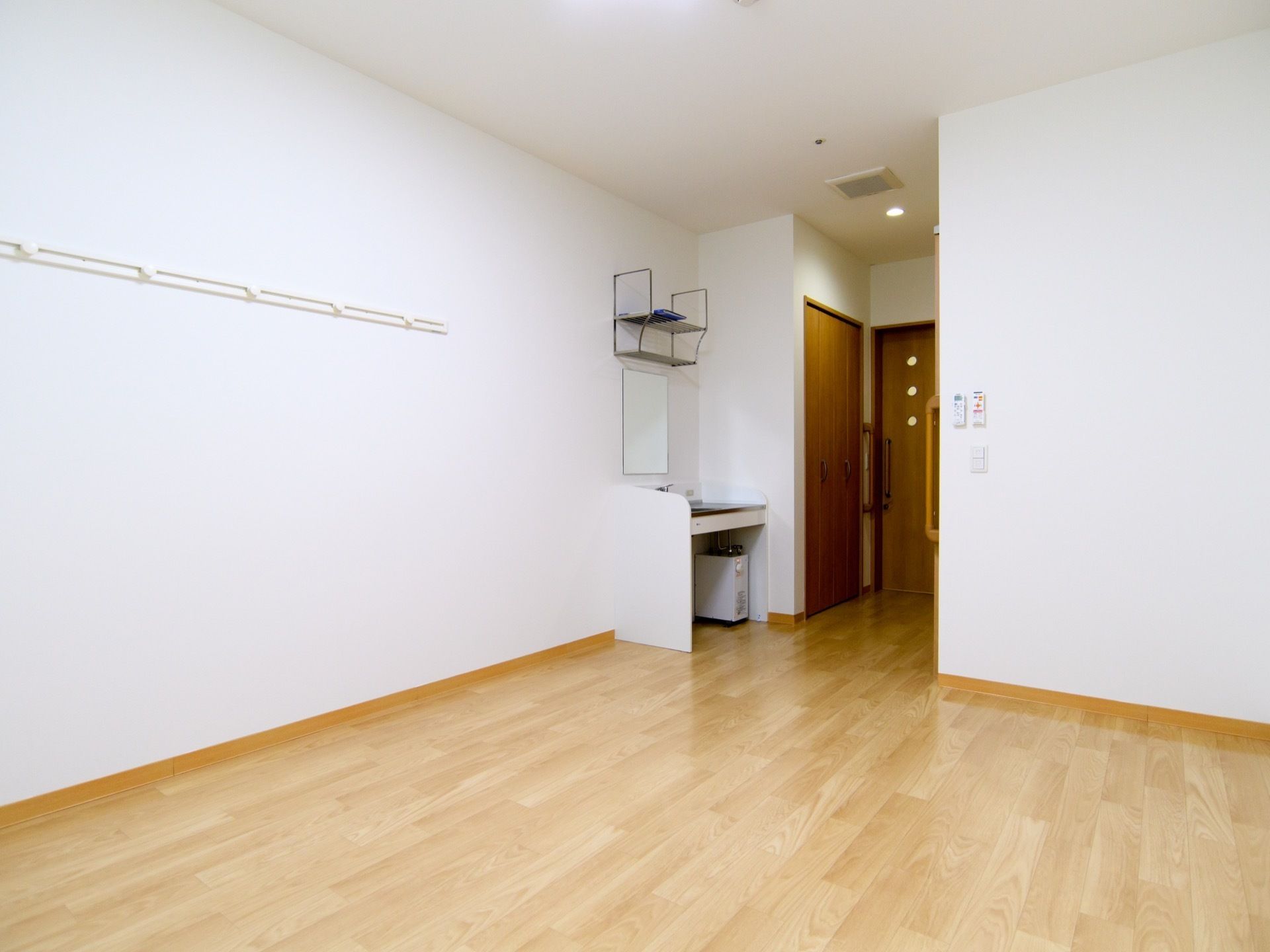 木目調のフローリングの居室である。部屋の一角に洗面台を備えている。壁には、白の壁掛けフックを取付けている。