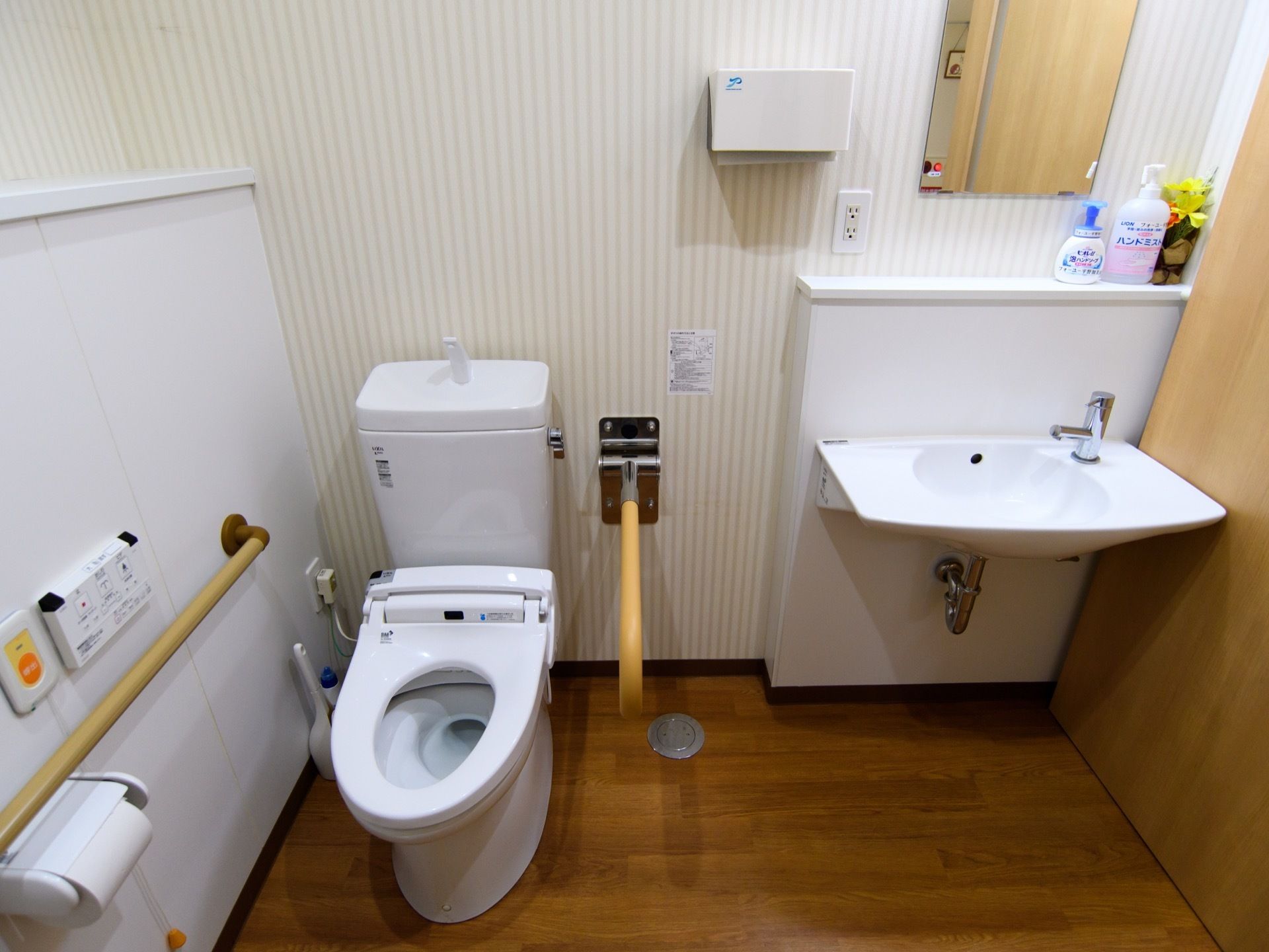 ストライプの壁に便器と洗面台を設置したトイレスペースである。便座の横側の壁に手すり、ナースコールなどを完備している。上部は棚になっている。
