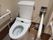 洋式トイレの左右に手すりがついている。壁に緊急時呼び出しブザーがある。床に段ボールがあり、新聞紙が置いてある。