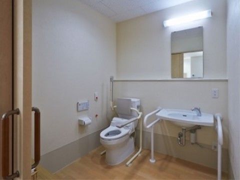 便器にはトイレ用の肘掛けが設置されている。同じ空間に手洗い場がある。壁にはスタッフを呼ぶためのボタンが設置されている。