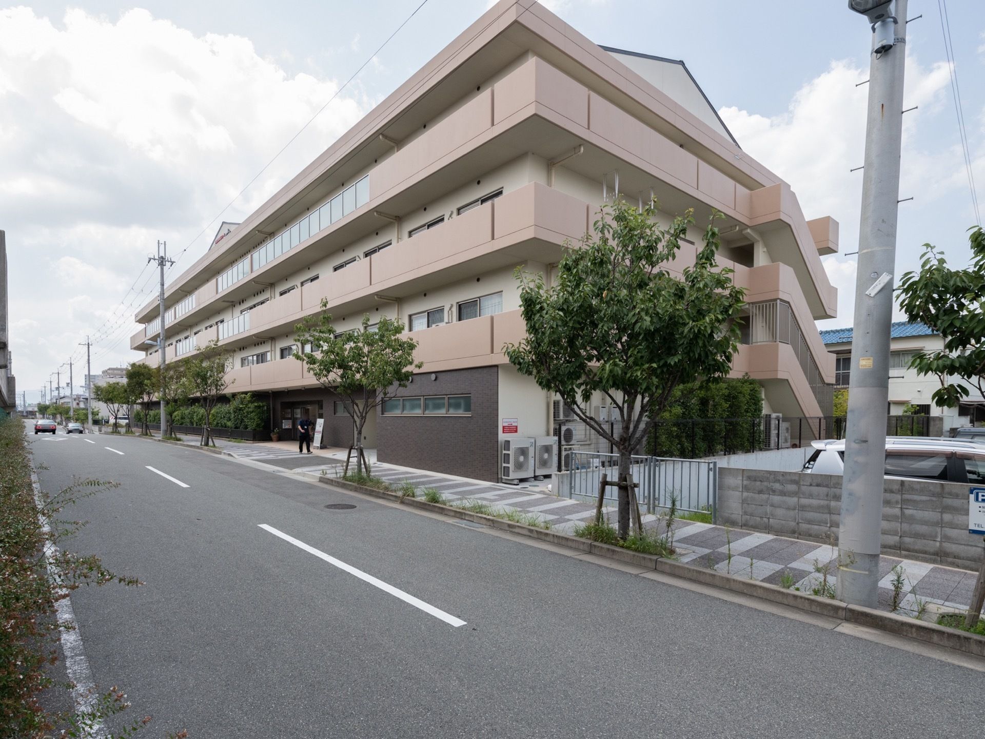施設の前には等間隔で木が植えられており、すぐそばは道路になっている。４階建てでベージュ色の外観である。