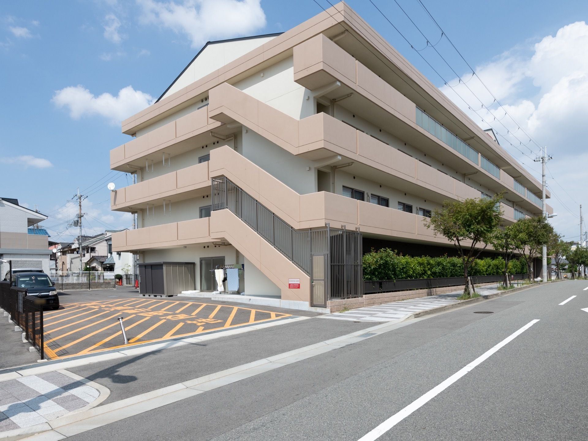 施設は４階建てで１階部分は植え込みの緑で囲まれている。白色とベージュ色の外観で、外階段は鍵付きになっている。