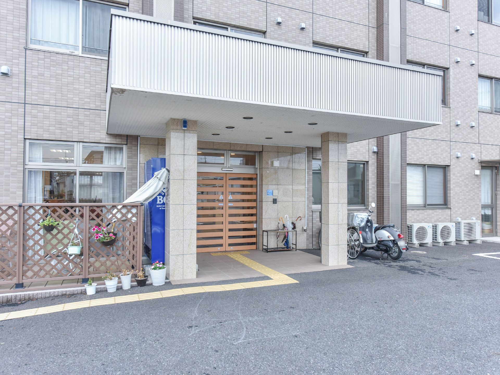 正面玄関脇には植木鉢のお花が飾られている。玄関へと続く点字ブロックを設置している。自動販売機、駐輪場、傘立てがある。