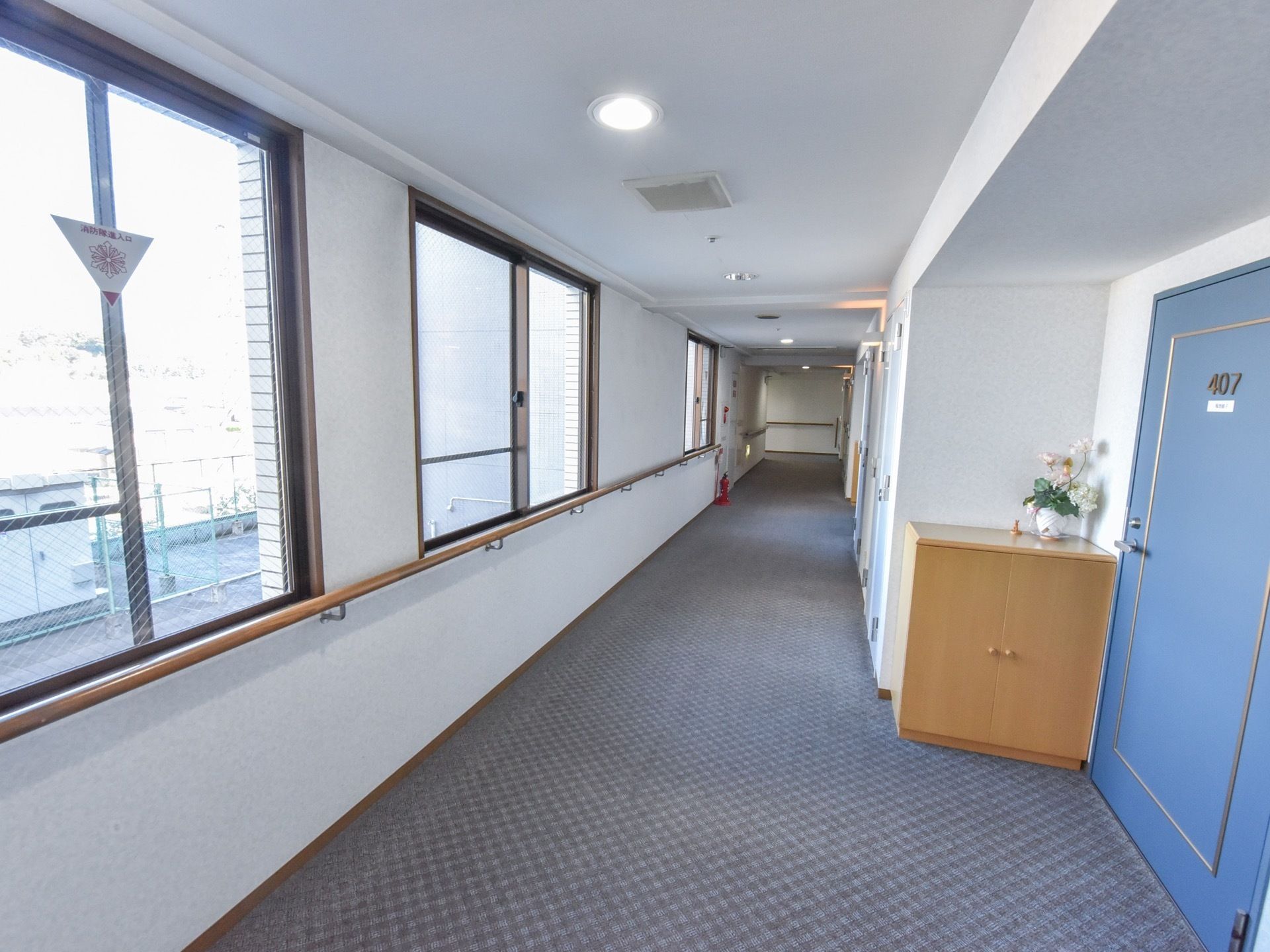 カーペットの敷かれた廊下は壁際に手すりが設置されており、入居者様の居室につながる青いドアも確認できる。