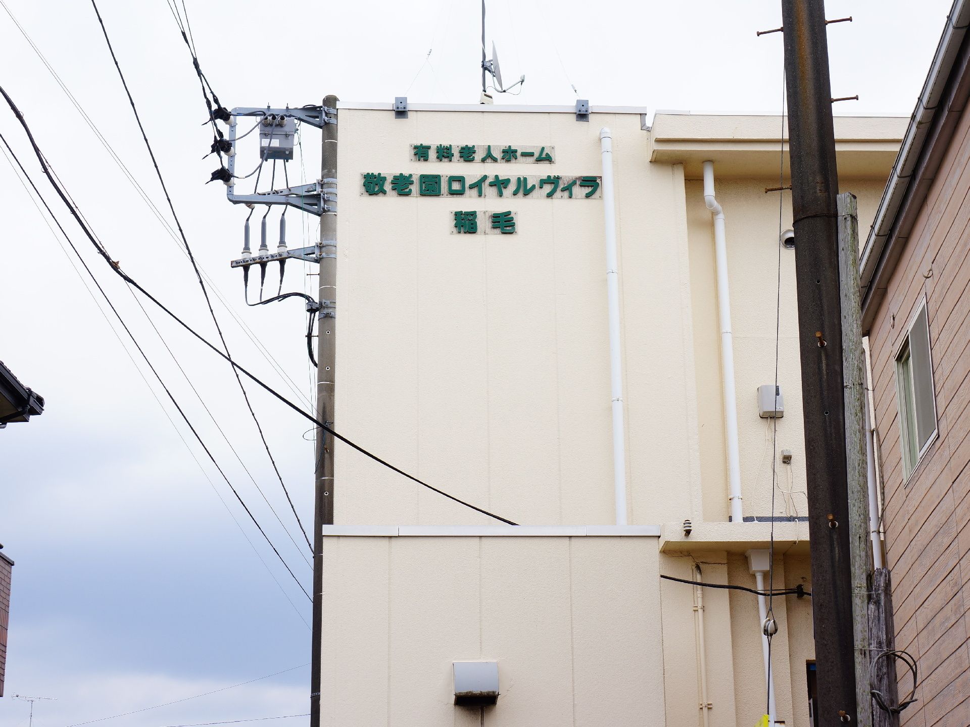 ベージュ系の建物の2階部分に、緑色の文字で施設名が表示されている。その文字は目立つ。建物は住宅街の中に建っている。
