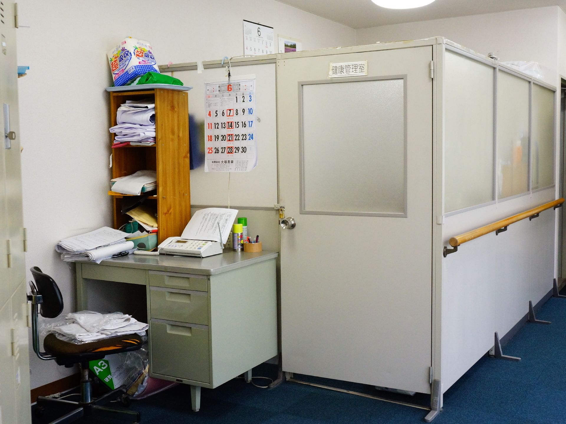 健康管理室のドアの横には、デスクがある。デスクの上には電話があり、棚にはものがたくさん置かれている。