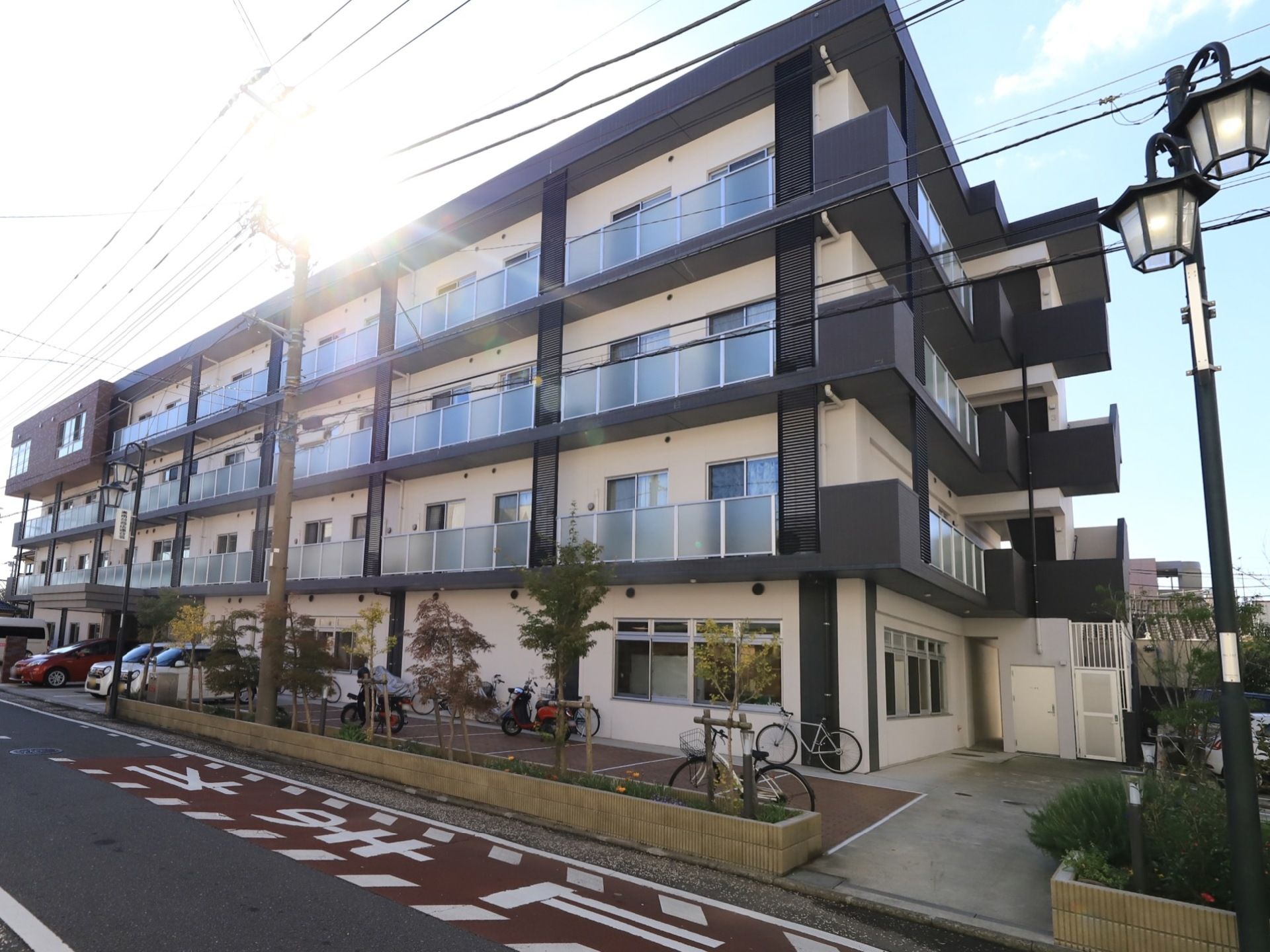 道路沿いの敷地に、モノトーンの４階建てマンションが建っている。建物の横には駐車場と駐輪場が設けられている。