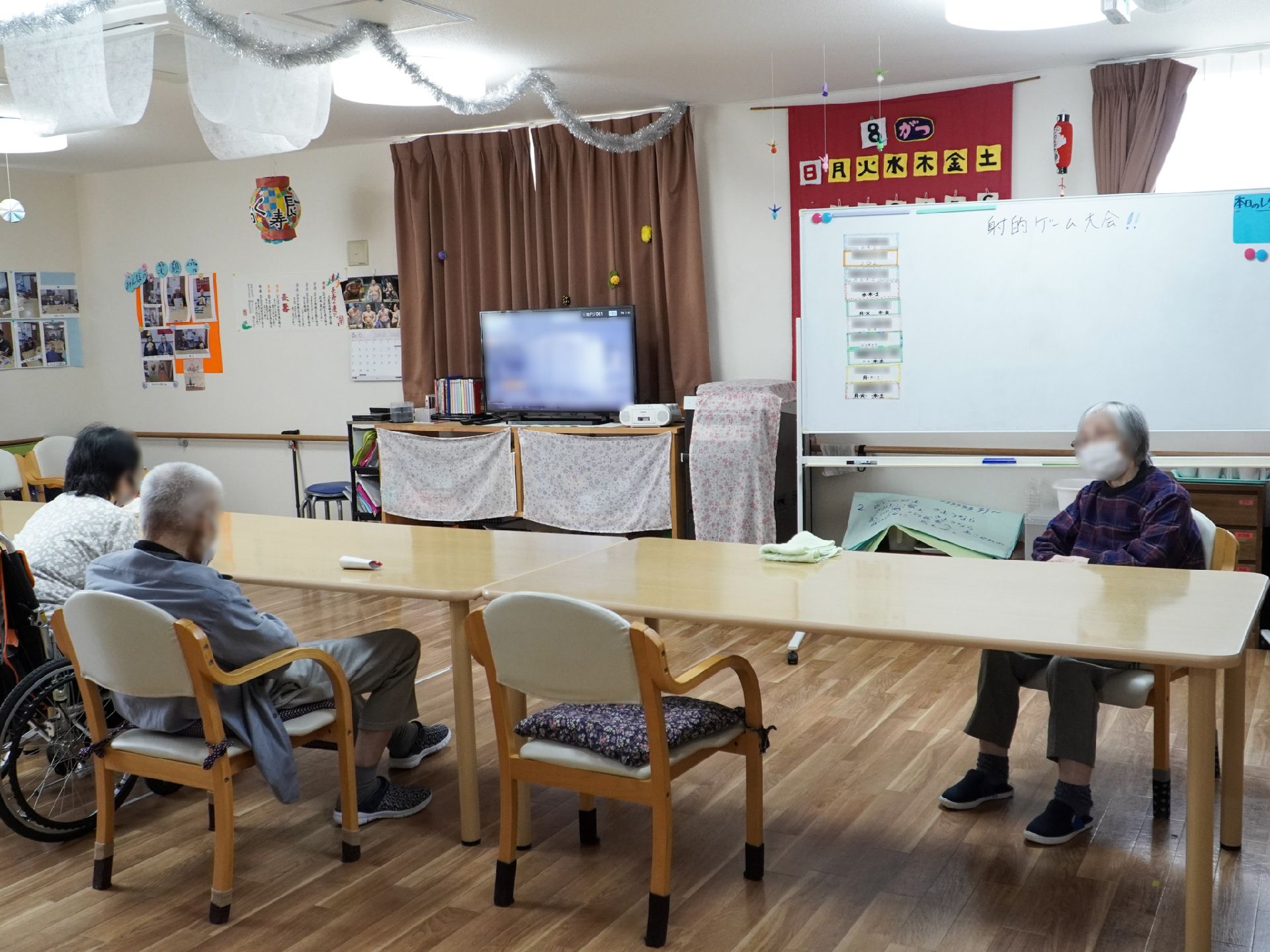 白い壁の広間に置かれた木製の長机の周りに集まる入居者様