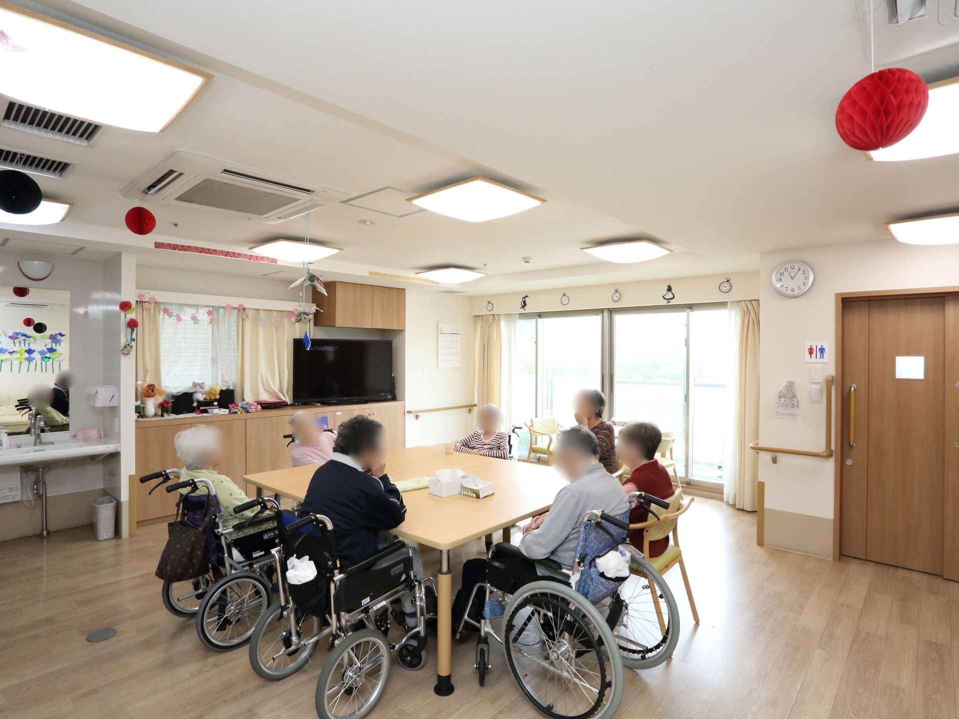 入居者様が大きなテーブルを取り囲み談話している。テーブルから見やすい位置にテレビがあり、奥のバルコニーへ続く窓からは陽が差し込んでいる。