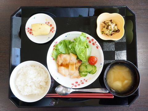 黒いお盆の上にはスプーンとお箸が用意されている。ごはん、お味噌汁、レタスとキュウリなどの野菜、お肉、和え物、バナナが食事として提供される。