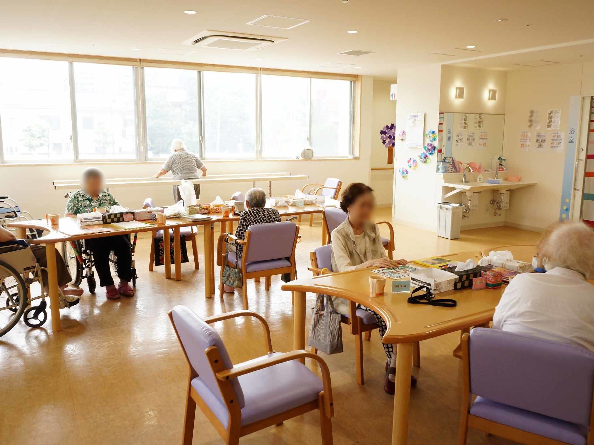 食堂には木の椅子とテーブルが並べられている。椅子や車いすに腰かけている入居者たちがいる。部屋の端にある平行棒を使っている方もいる。