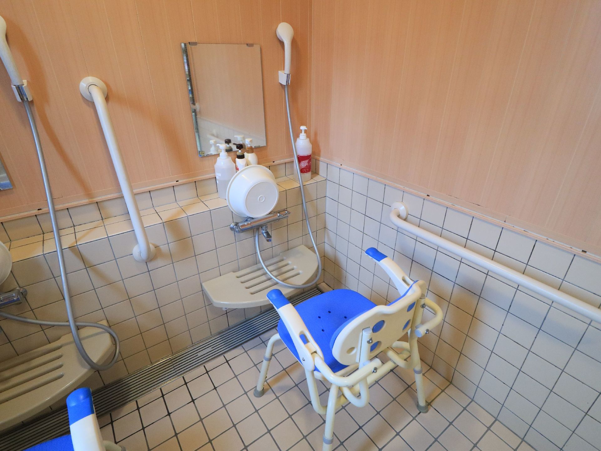 浴室に、可動式のひじ掛けがついた椅子が置かれている。洗い場の前には、ボディソープやシャンプーのボトルが並んでいる。