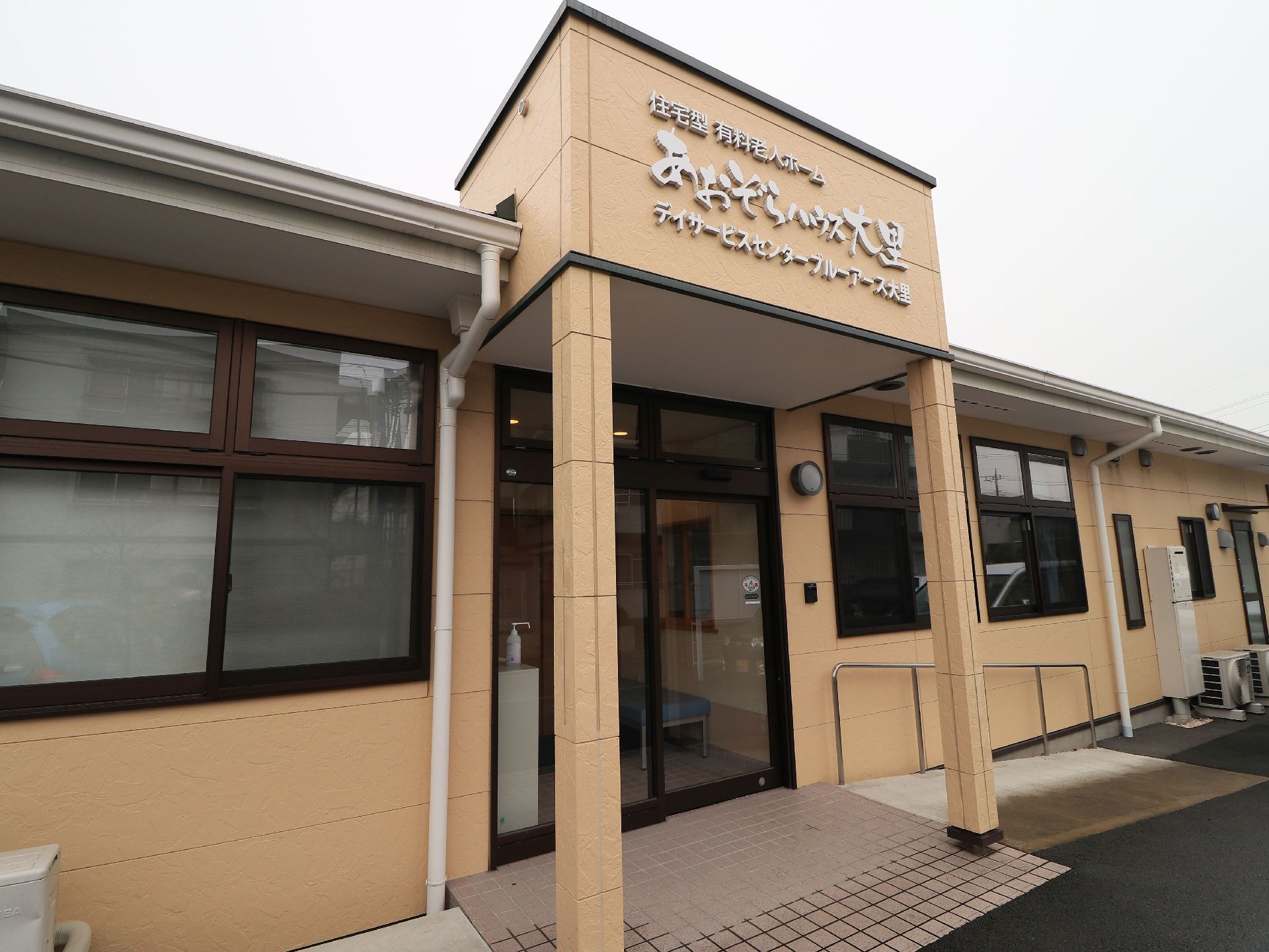 玄関の上部にひさしがついていて、そこに施設の名称が表示されている。玄関は黒枠がついたガラスの自動扉になっている。