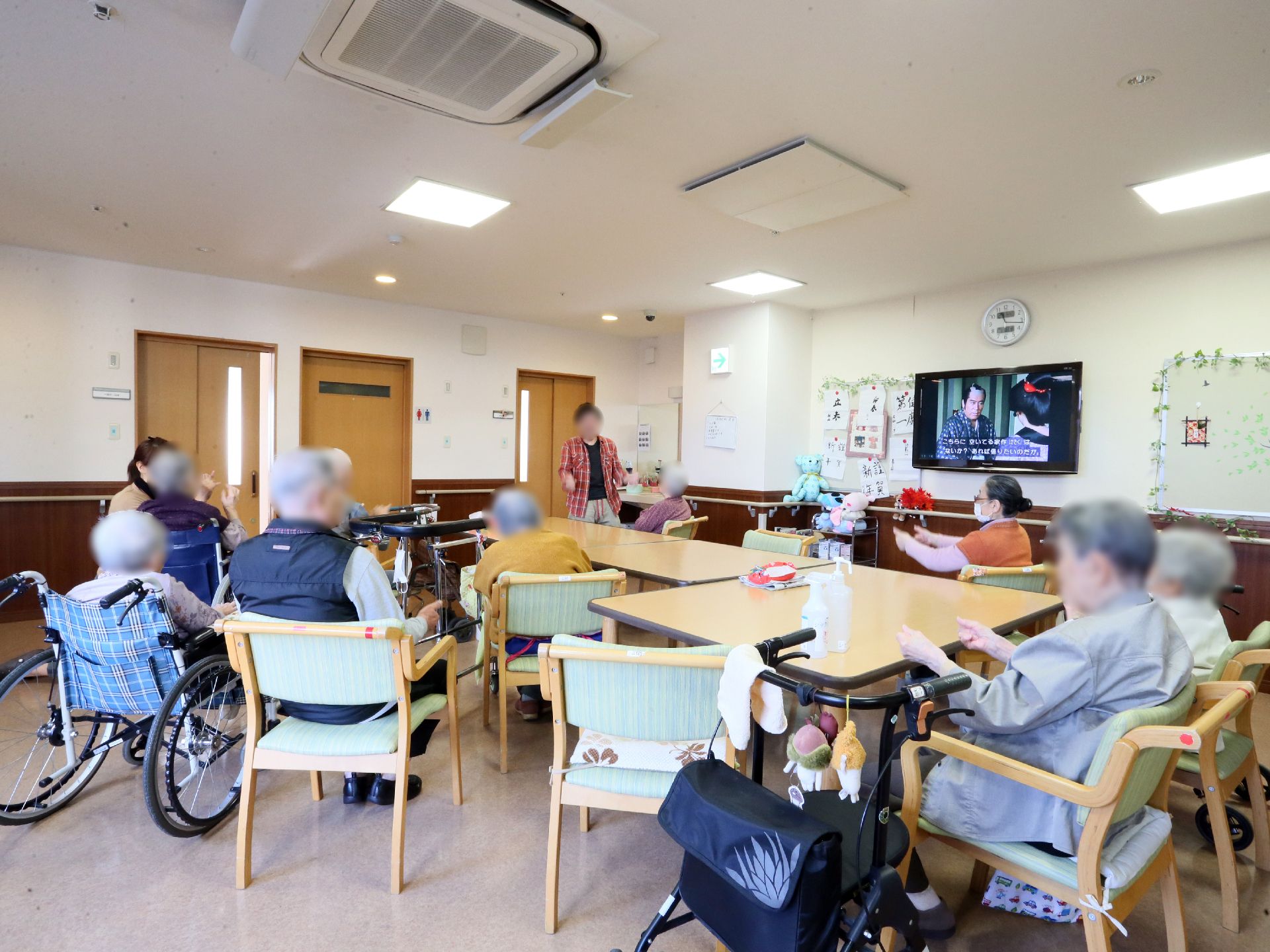 テーブルの周りに入居者が集まっている、壁にテレビがかけられた部屋