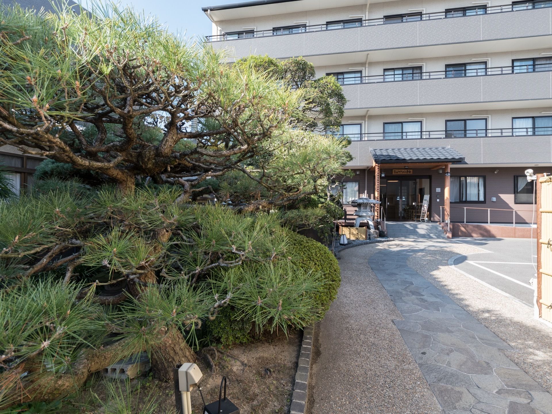 正面玄関へと続くアプローチの横には、松などの植木がある。正面には手すり付きの階段が、横にはスロープが備わっている。