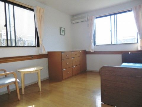 ２箇所に窓が設けられた明るい居室である。エアコンや介護ベッド、収納家具が備わっている。壁には、手すりが設置されている。