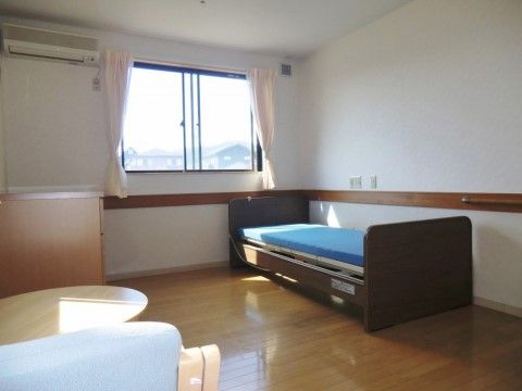 フローリングの居室は個室になっている。ナースコールや手すり、エアコンが完備されている。介護ベッドや収納家具が壁際に配置されている。