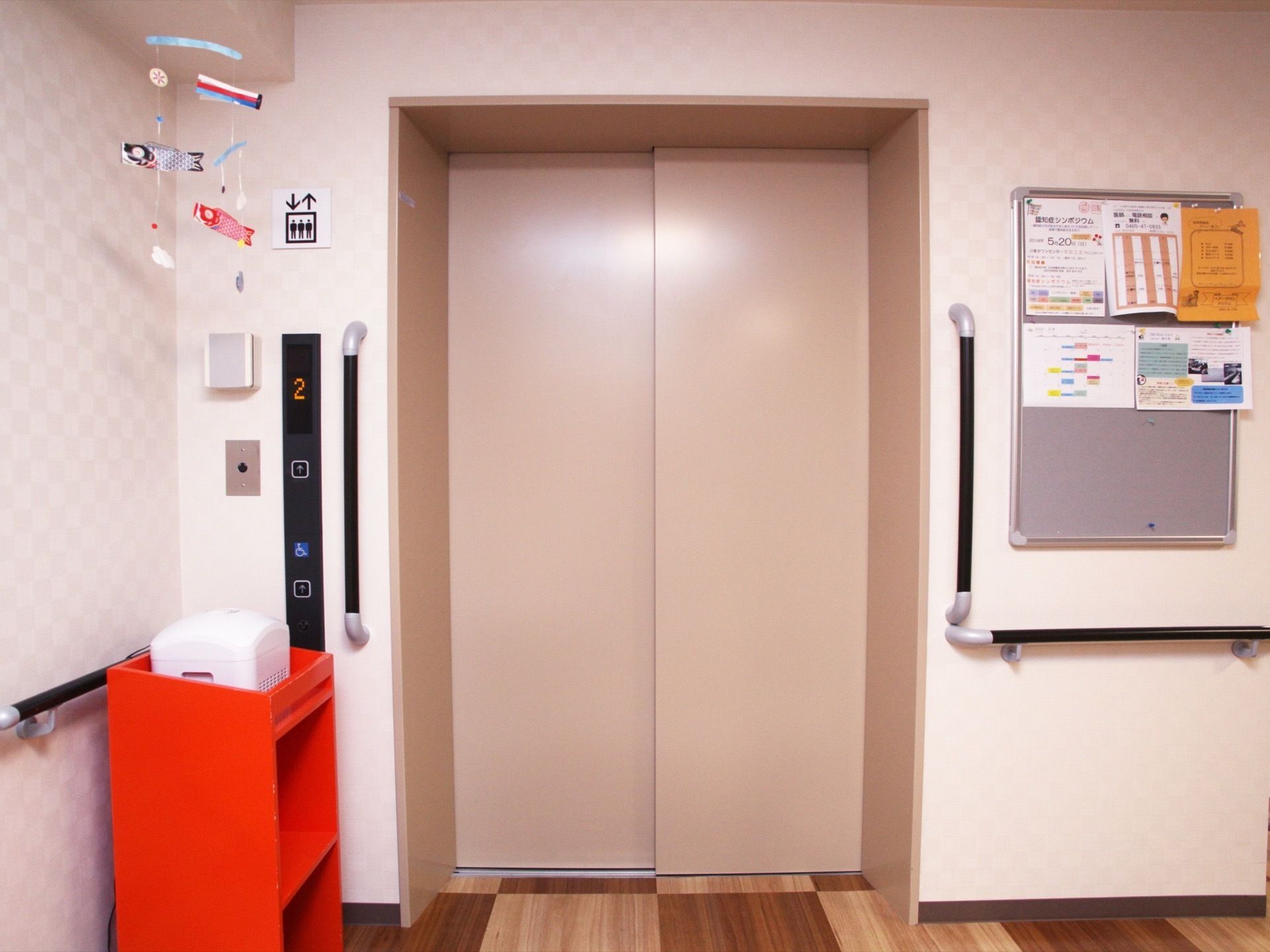 淡い柿色のエレベーターの扉の横に、掲示板が設置されている。掲示板には、お知らせのチラシなどが何枚か貼られている。
