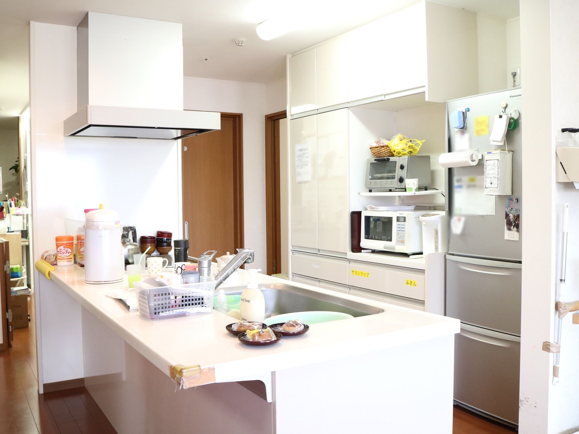 白色を基調とした対面式のシステムキッチンである。壁に沿って収納棚が設けられ、冷蔵庫や電子レンジ、トースターなどの電化製品が置かれている。