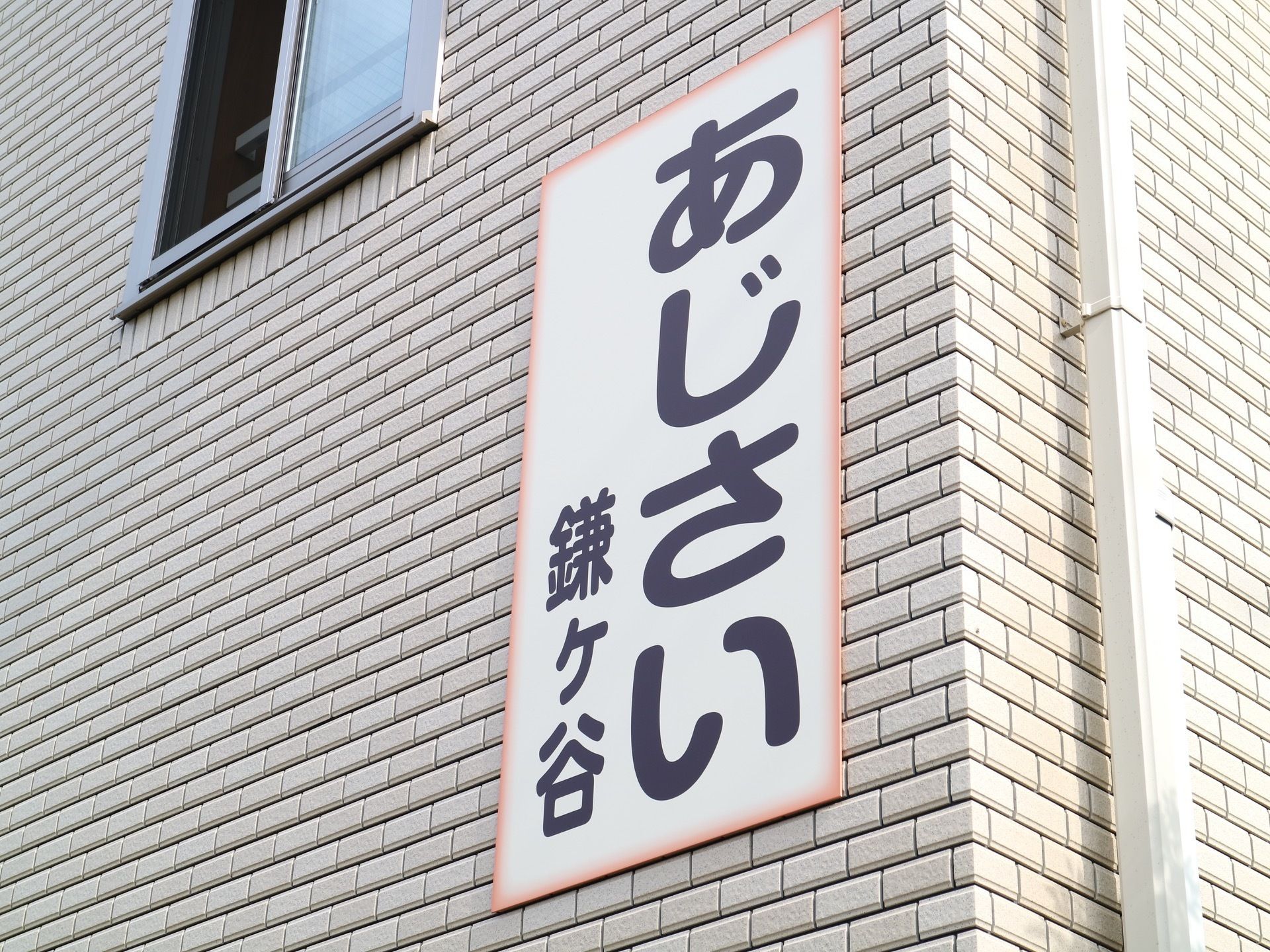 ベージュのタイル張りの壁に、白い看板を設置している。枠線はオレンジ色、文字は黒色の太い線で描かれている。