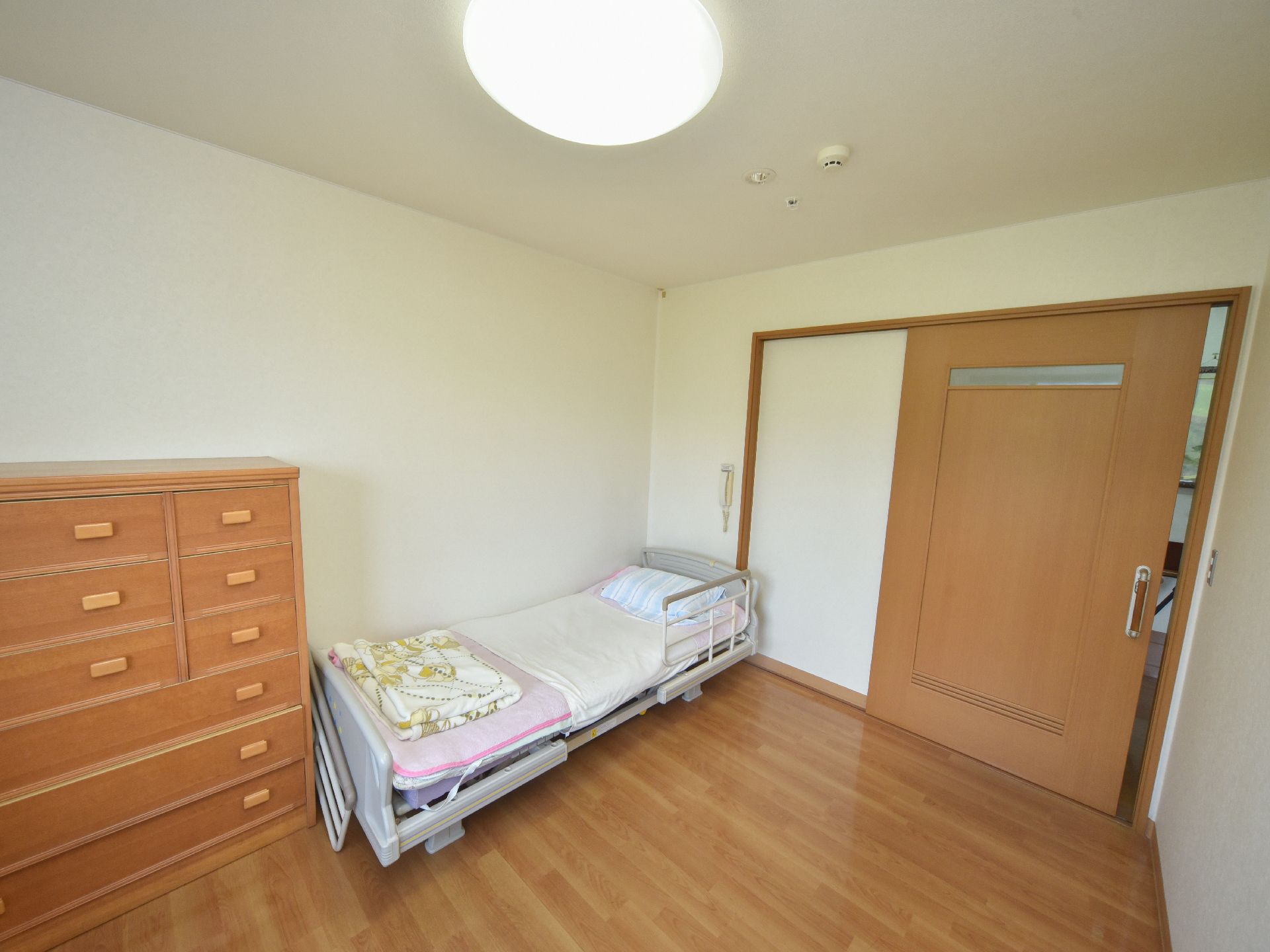大きな茶色い扉がある部屋は広々としている。その中に手すり付きのベッドと6段からなるタンスが1竿置かれている。