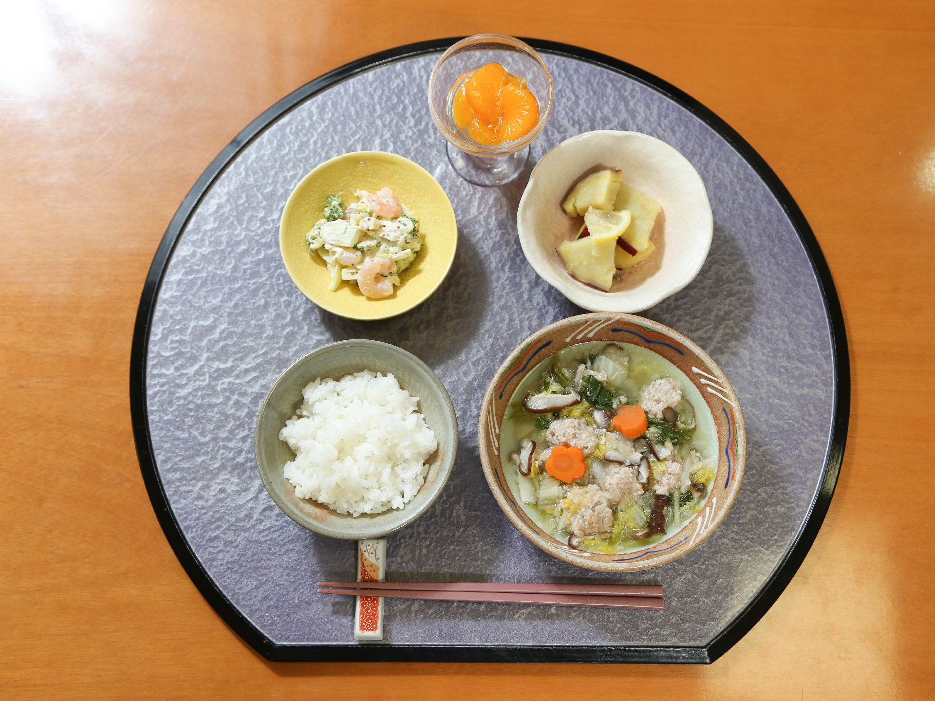 食事。白米に、肉団子、白菜、ニンジン、シイタケが入った澄まし汁、ブロッコリーとエビのサラダ、サツマイモ、デザートにみかんがある。
