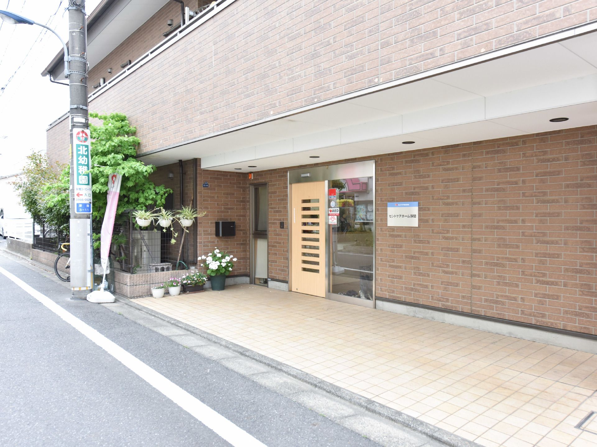 施設の前はアスファルト舗装の道路である。玄関前の床はタイル張りで、花が植えられている鉢が複数置かれている。