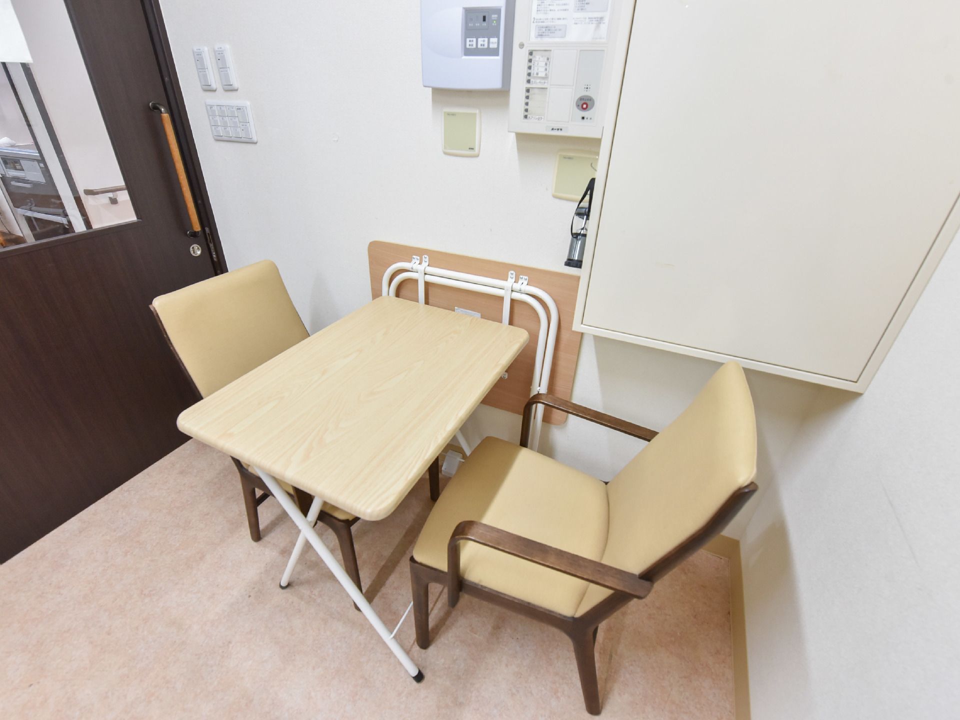 長方形の天板のテーブルに椅子が2脚セットされている。横には折り畳みテーブルが立て掛けられている。床はカーペット敷きである。