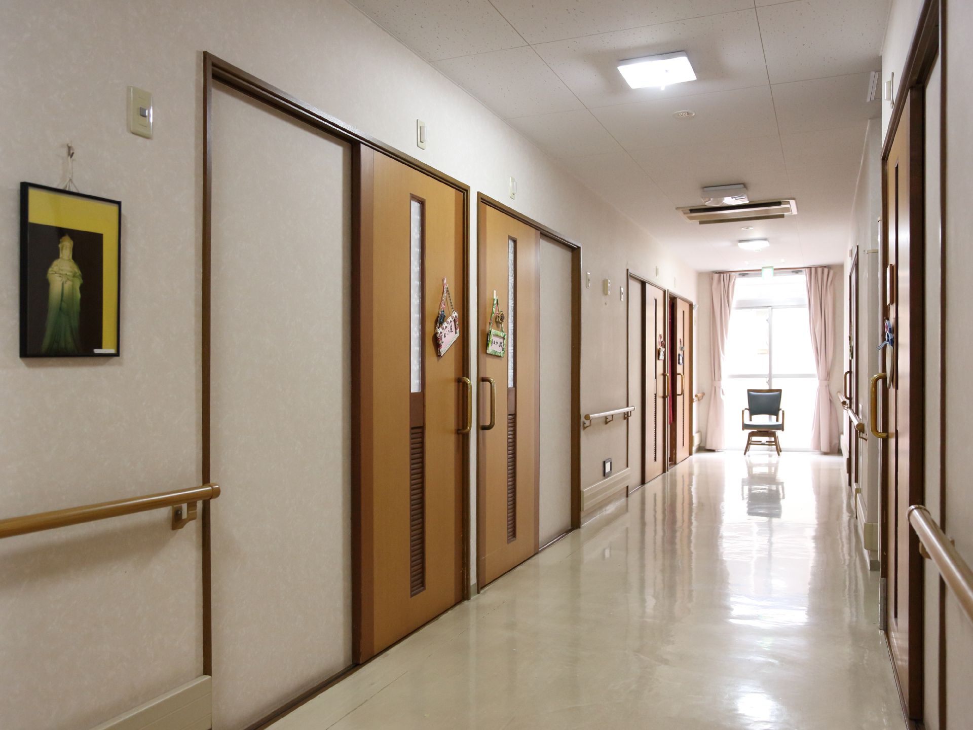 左右に部屋のドアが並んだ廊下。突き当りは天地一杯の窓になっており、手前に椅子が置かれている。壁には絵が掛かっている。