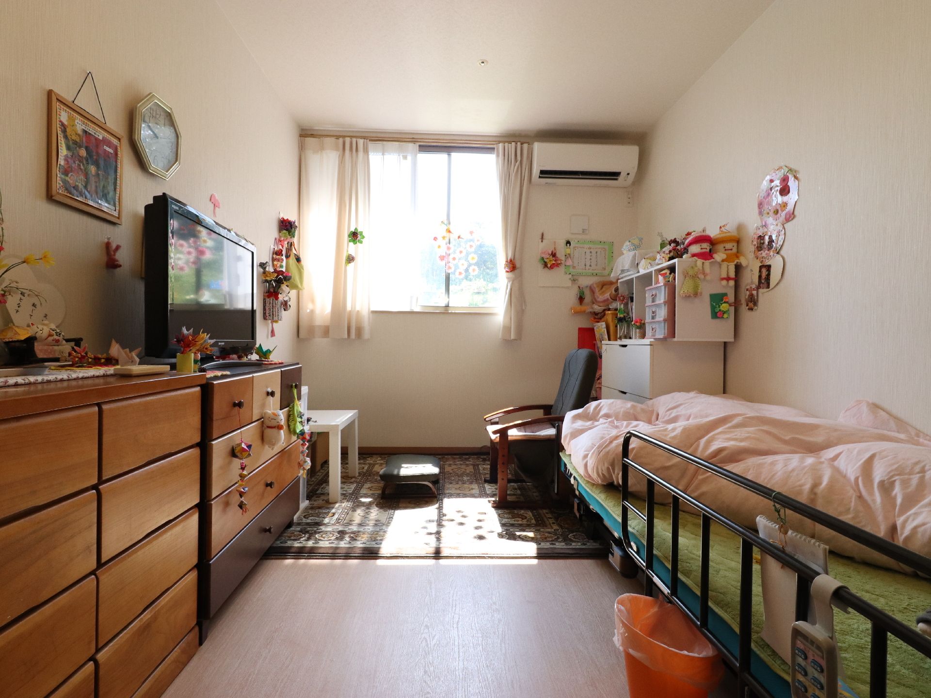 居室の窓からは日光が差し込み明るい。テーブル、椅子、テレビ、タンス、クーラー、介護用ベッドが備わっている。