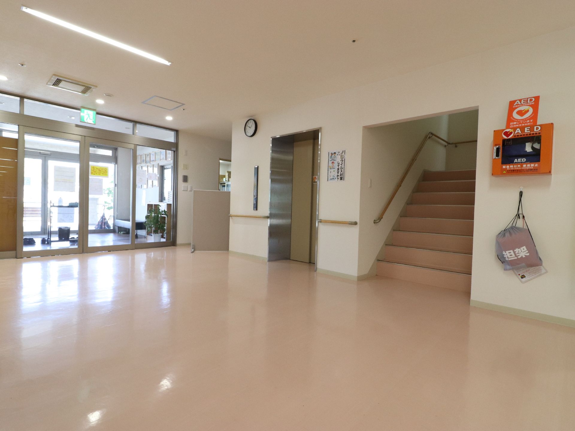 入口を入るとエレベーターと階段がある。壁には、AEDが設置されており、担架もかかっている。エレベーター上には、時計も設置してある。
