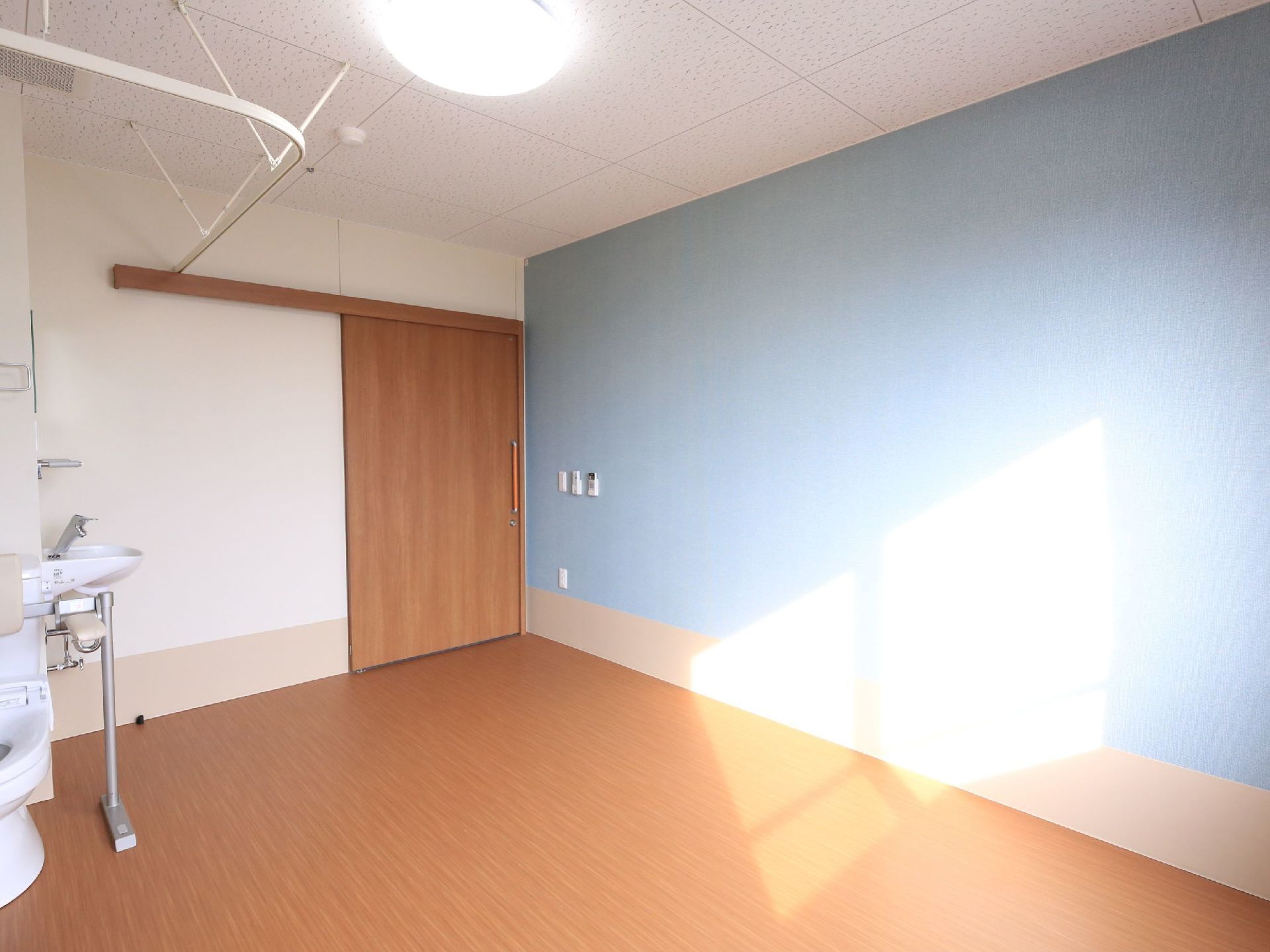 ロイヤルブルーの壁が印象的な広めの間取りの居室である。トイレや洗面台、照明が完備されている。仕切り用のカーテンが設置されている。
