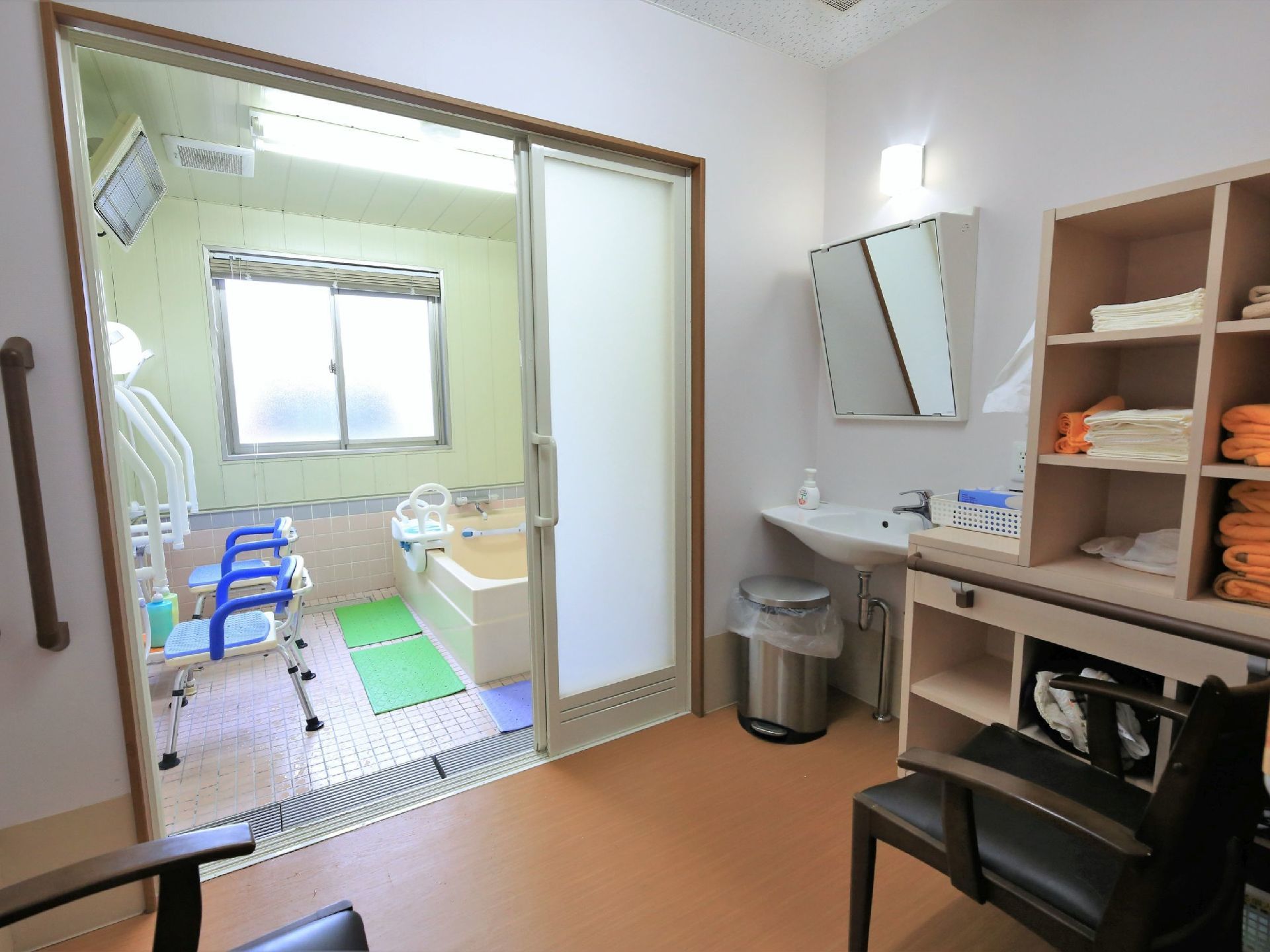 介護浴室の脱衣所である。壁際には、タオルが置かれた収納棚や車椅子対応洗面台が設置されている。椅子が置かれている。