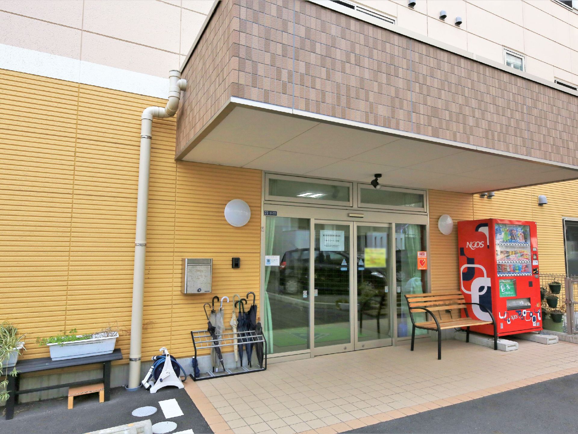 大きなひさし付きのエントランスである。自販機やベンチ、傘立てが出入口の自動ドア近くに設置されている。