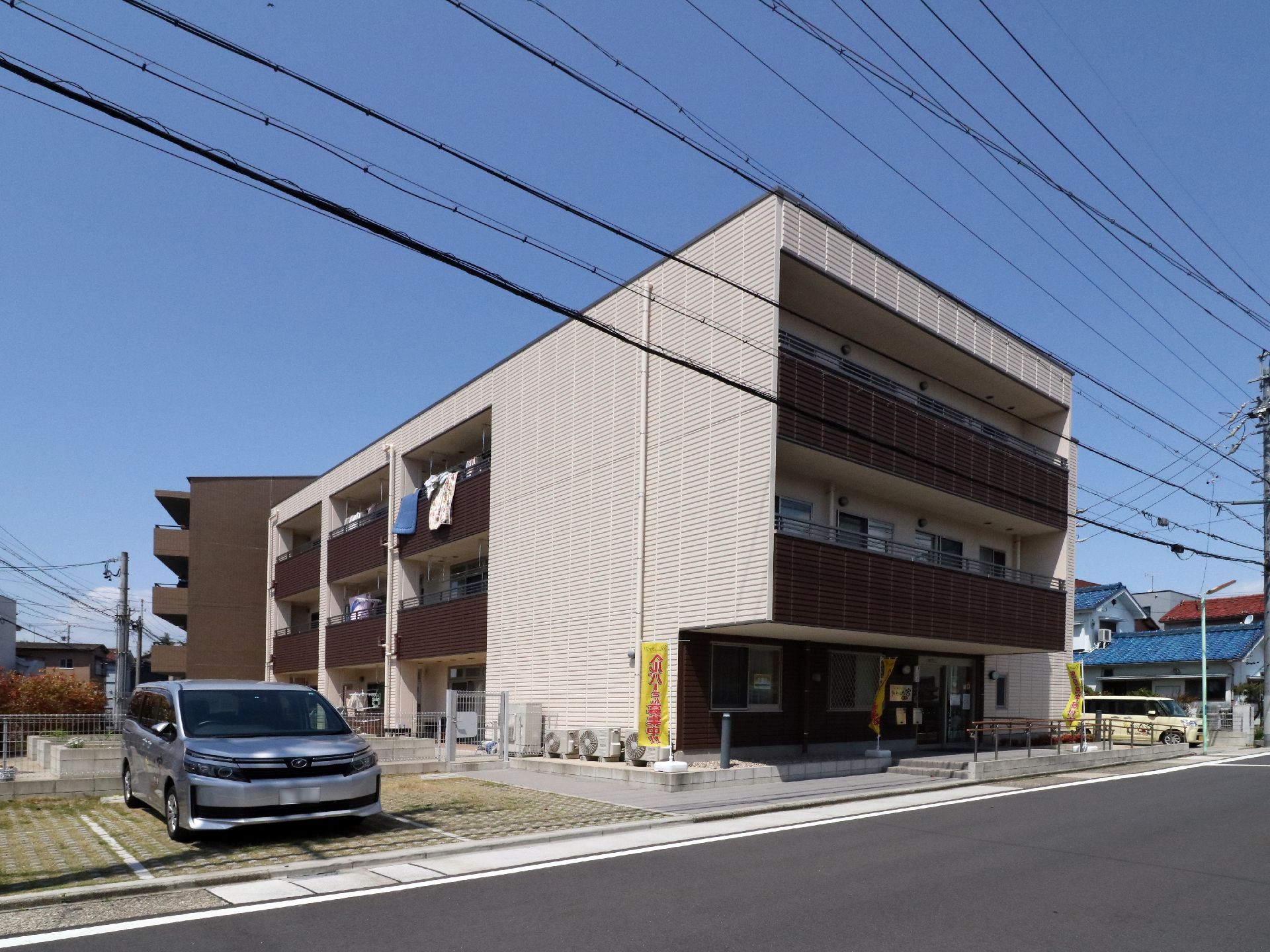 新しく舗装された道路に面した、ヘルパーを募集するのぼりが立つ３階建ての施設