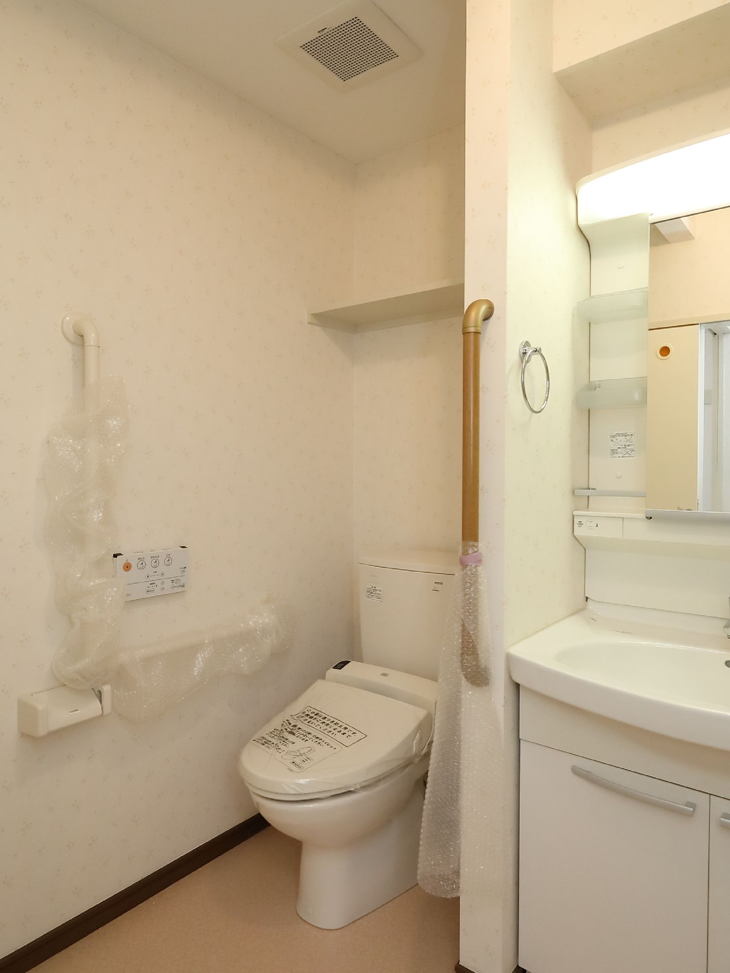 白を基調としたトイレで、左奥に温水洗浄便座がある。操作用パネルは壁に取りつけられている。壁で仕切られ、洗面台がある。