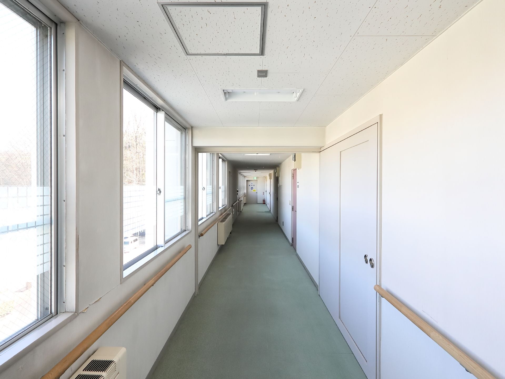 廊下の左手は一面窓になっている。窓の下に手すりが取りつけられている。壁には固定型のヒーターが設置されている。