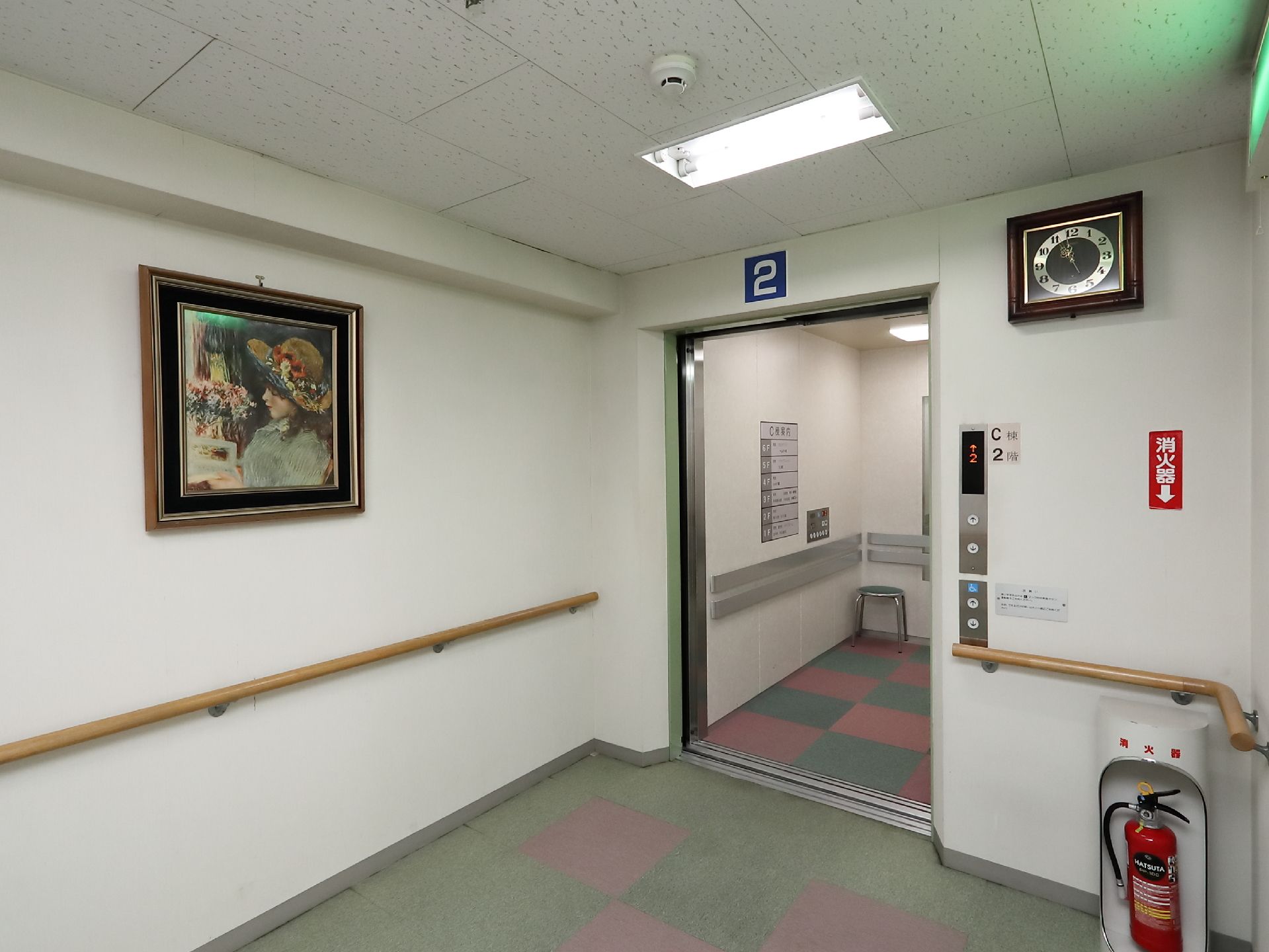 エレベーターホールの壁に大きな絵画が飾られている。エレベーターは開いており、扉の右側に呼び出しボタンがある。