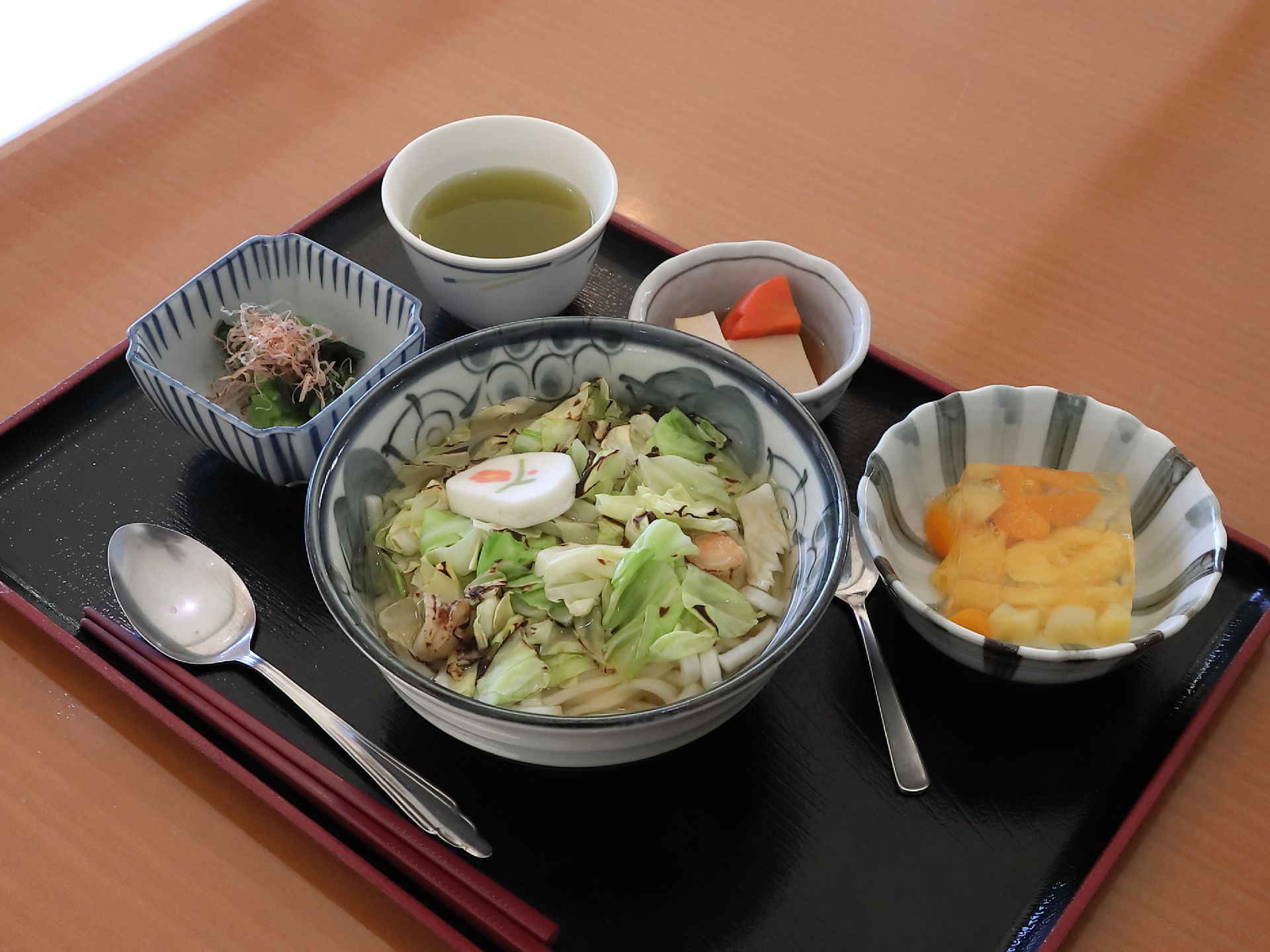 食事は１人分ずつトレイに乗せられて配膳される。野菜がたくさん乗ったうどんが主食で、小鉢やデザートがついている。