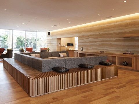 壁一面が窓で明るい。中央にコの字型の大勢が座れるソファがあり、クッションが並ぶ。窓際にはソファセットが並んでいる。