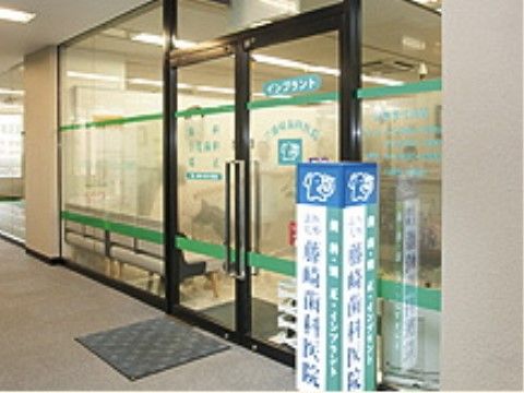 駅近くのビル内にある歯科医院。インプラント治療も行う。入り口はガラスの扉で観音開き。待合室にはソファが並んでいる。