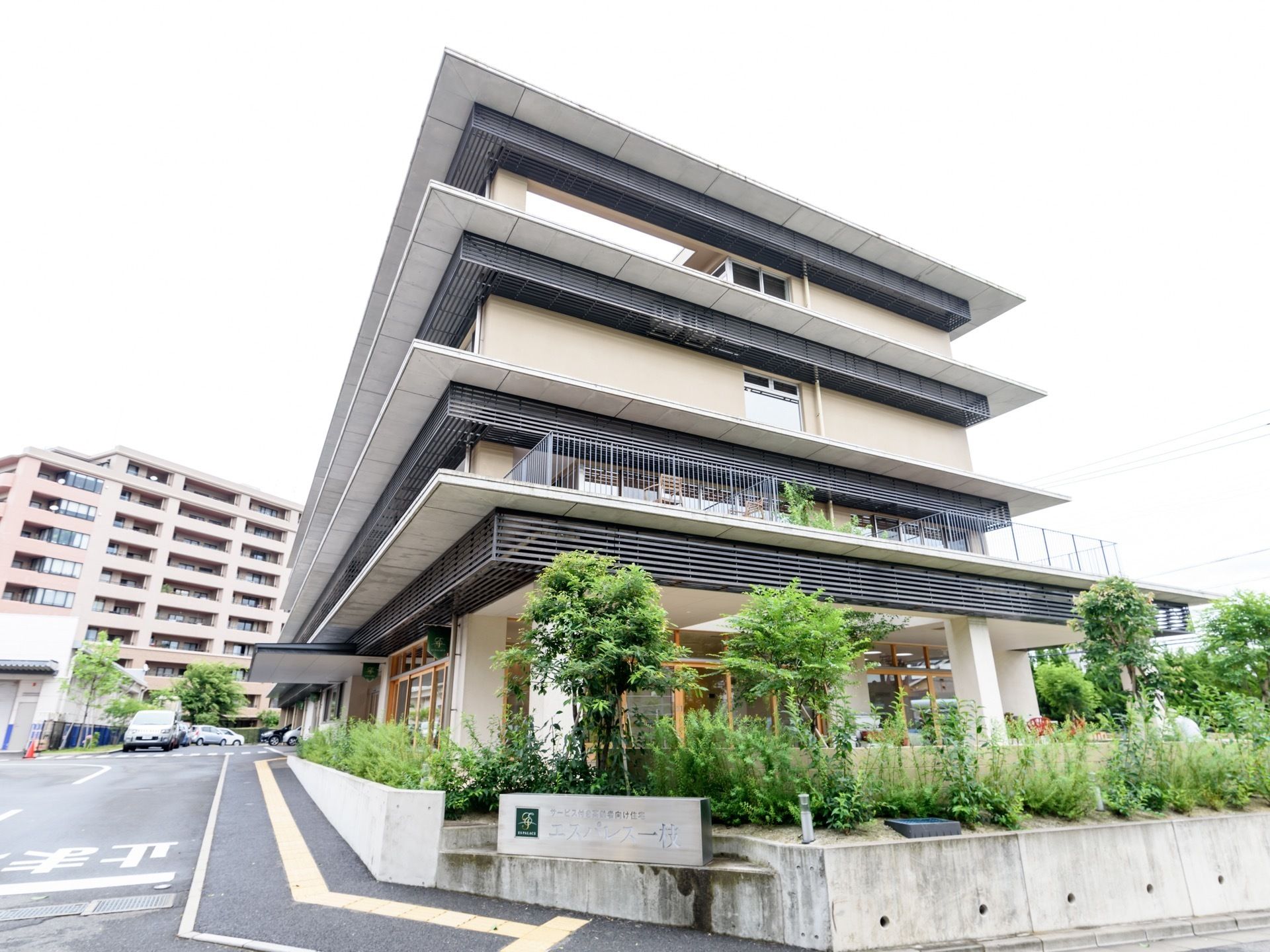 道路側から施設を見ると、手前には整備された植栽があり、緑の溢れた空間にスマートでモダンな建物が建っている。