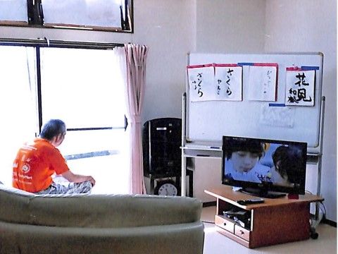 ソファに腰を掛けた入居者様がテレビを見ている。テレビの向こう側にはホワイトボードがあり、入居者様の書道作品が飾られている。
