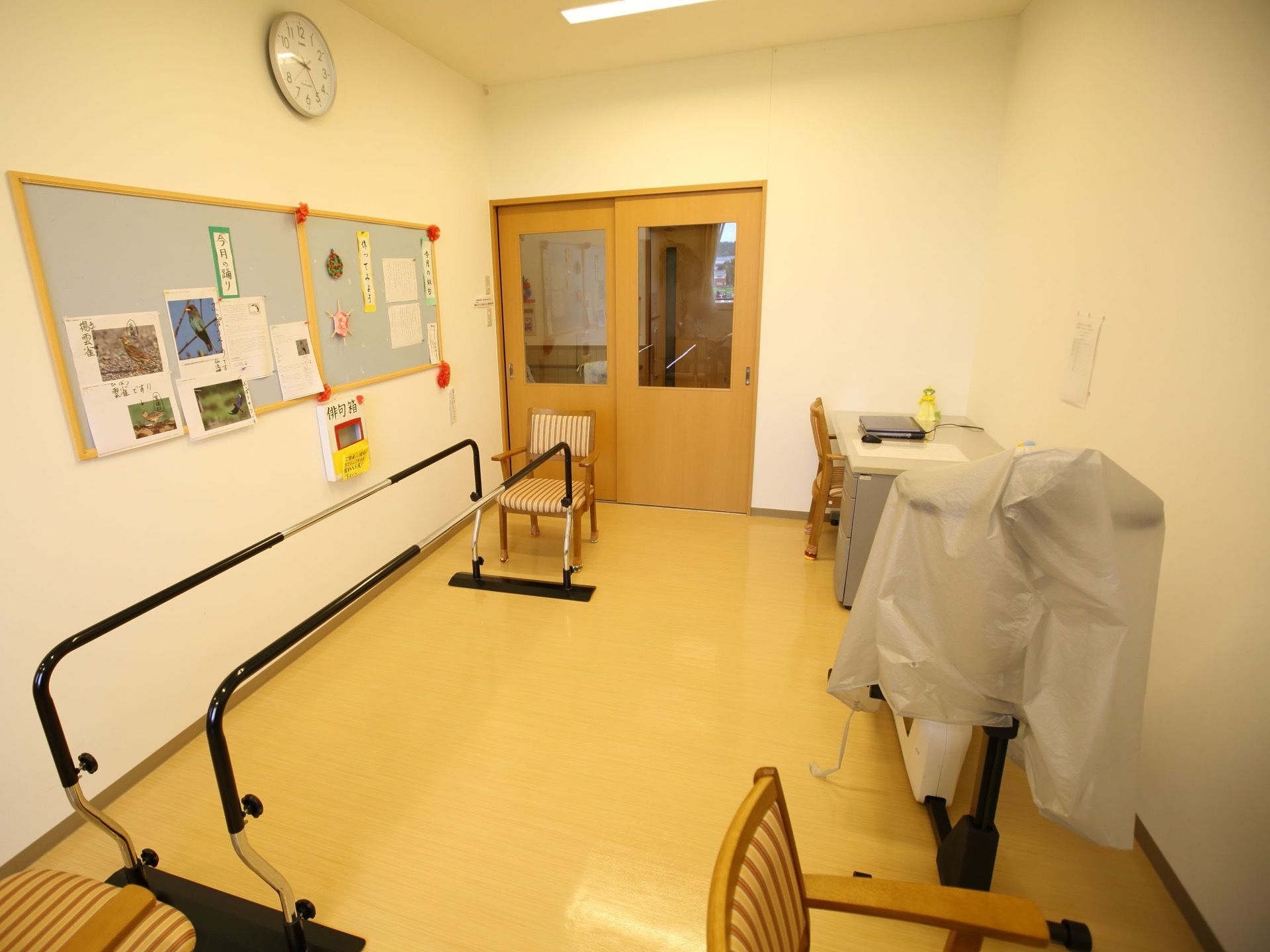 フローリングの床の上に椅子と歩行訓練用補助器具が置かれている。壁には入居者様の作品が展示されている。