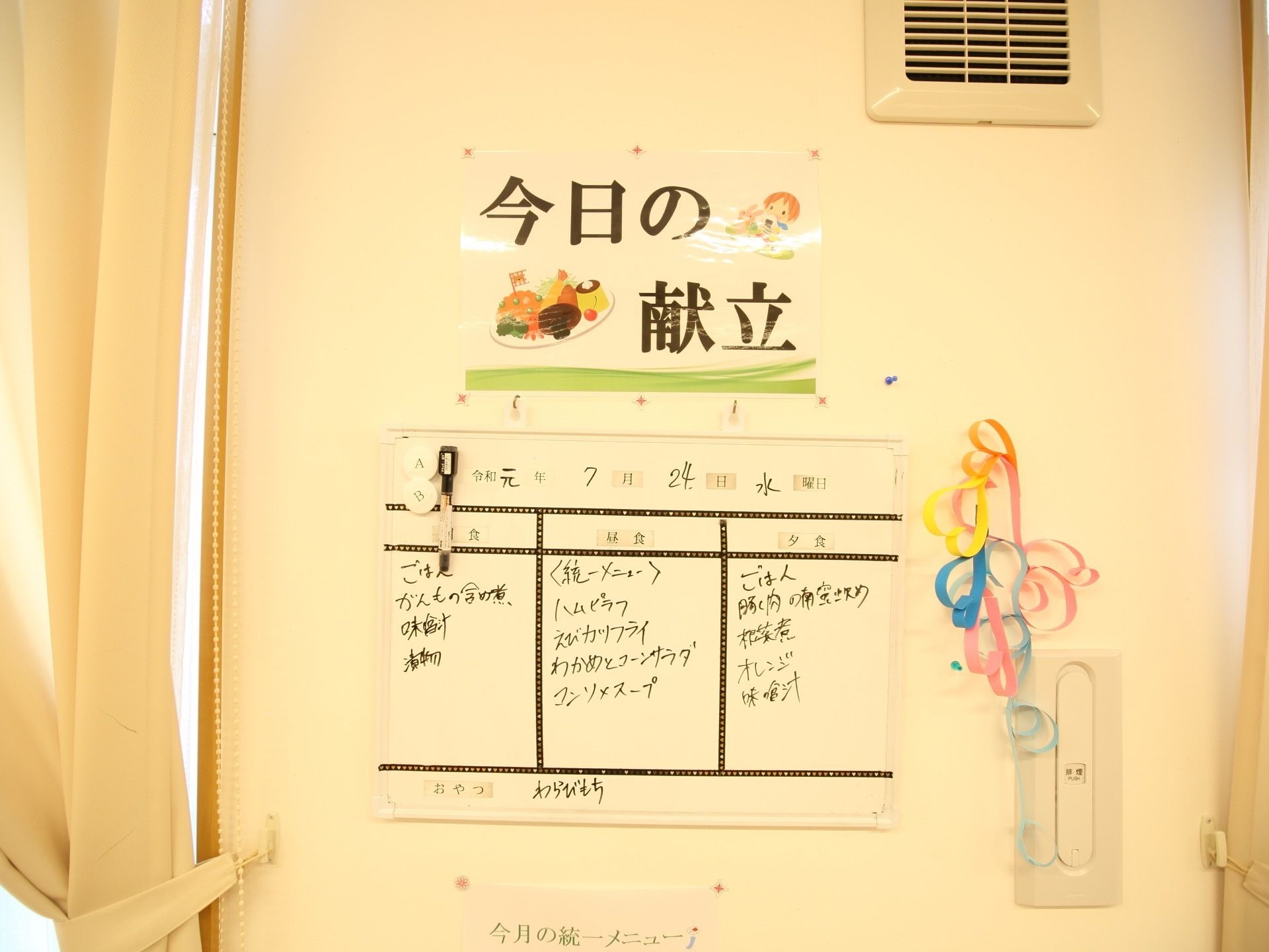 窓の横にあるスペースにホワイトボードが設置されており、黒いペンで朝食、昼食、夕食、おやつの献立が書かれている。
