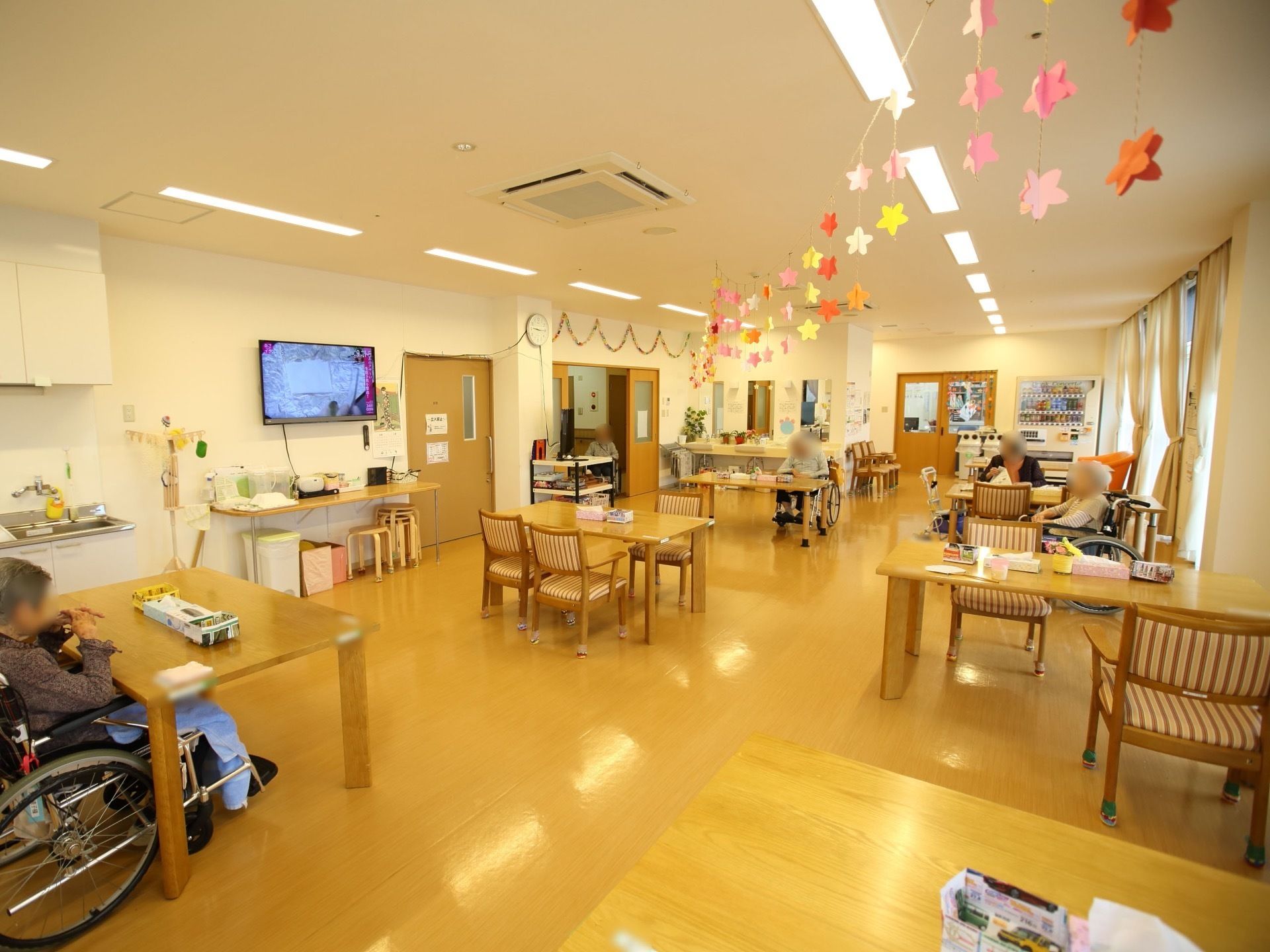 広々とした共有スペースにダイニングテーブルと椅子が置かれており、入居者様が座っている。壁掛けテレビが設置されている。