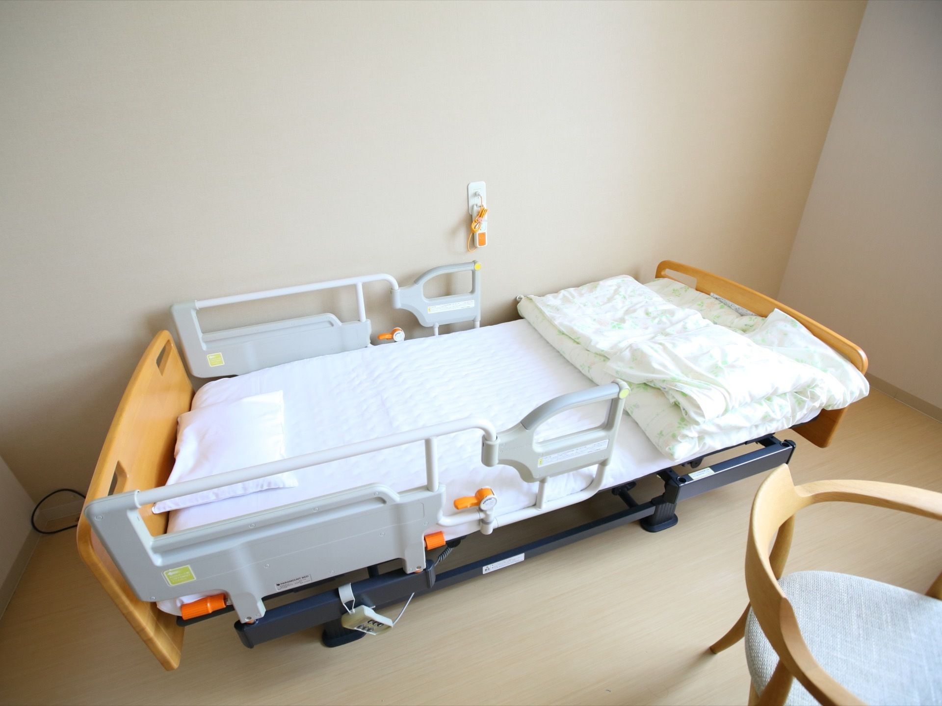 居室はベージュを基調としている。中央に介護用ベッドが置かれ、布団が敷かれている。壁にナースコールが設置されている。